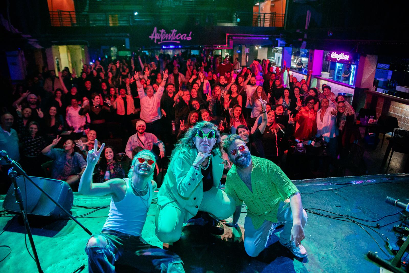 Três artistas agachados na beira do palco posam para a foto com o público d'A Autêntica ao fundo, muita gente de braço erguido.