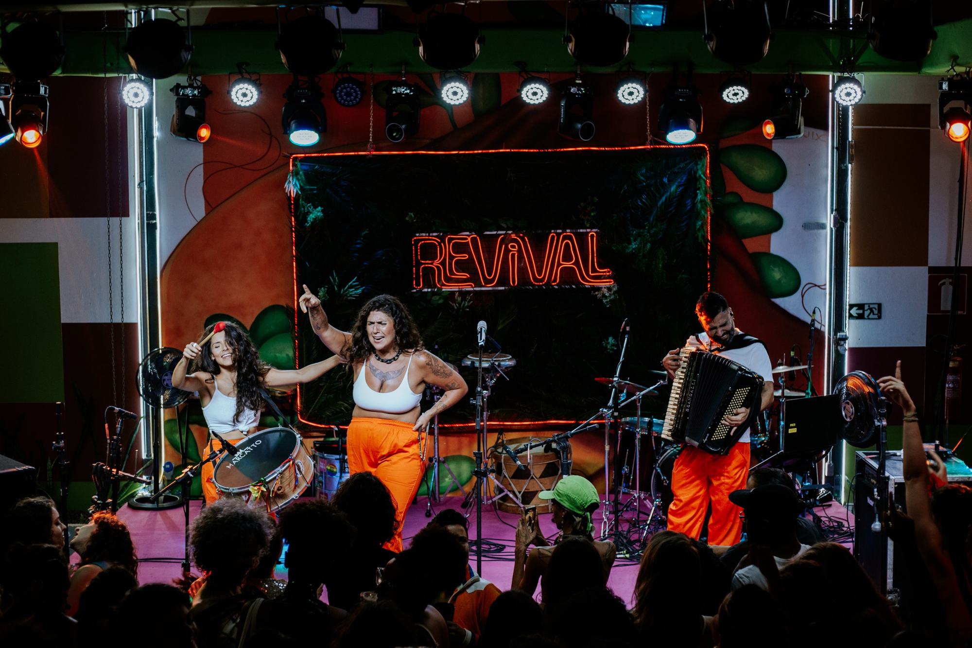 Cantora no palco, de top branco e calça laranja, com o braço erguido, entre uma percussionista e um sanfoneiro, diante do público.