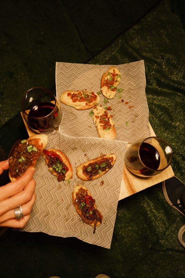 Bruschettas servidas sobre papel e duas taças de vinho tinto, com mãos à mesa.