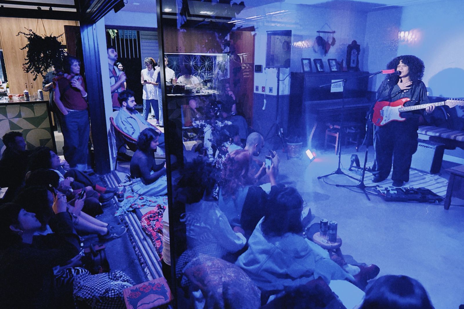 Show intimista numa sala com piano: a artista canta e o público assiste sentado no chão, sob luz azul.