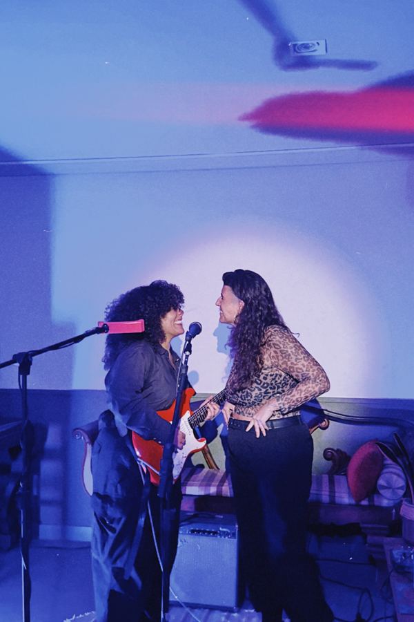 Duas artistas cantando juntas ao microfone, sob luz azul.