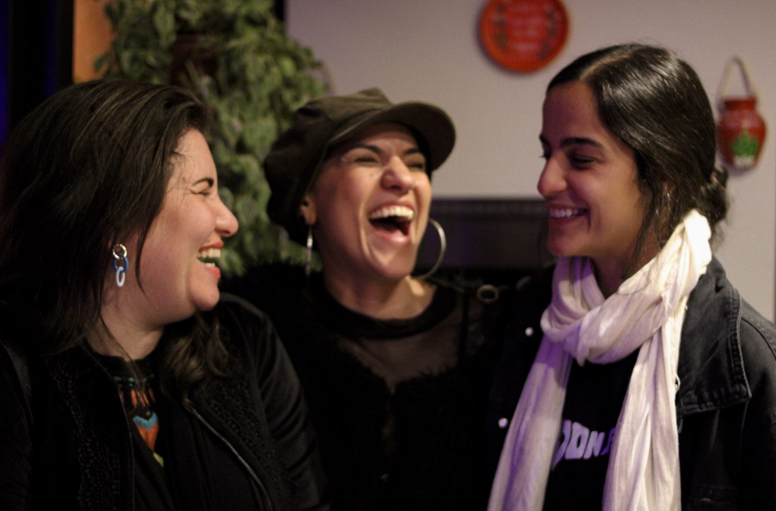 Três mulheres rindo juntas num ambiente interno, com plantas e um prato decorado na parede ao fundo.