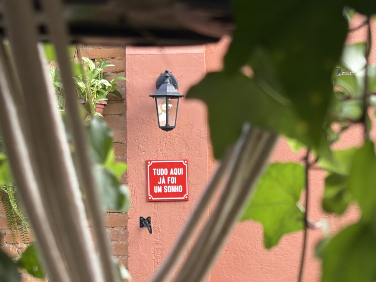 Placa vermelha com a frase “Tudo aqui já foi um sonho” numa parede rosada, vista entre folhas e o tecido de uma rede.