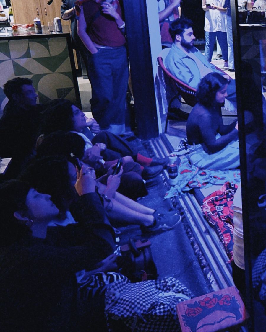 Público sentado no chão assistindo ao show, sob luz azul.